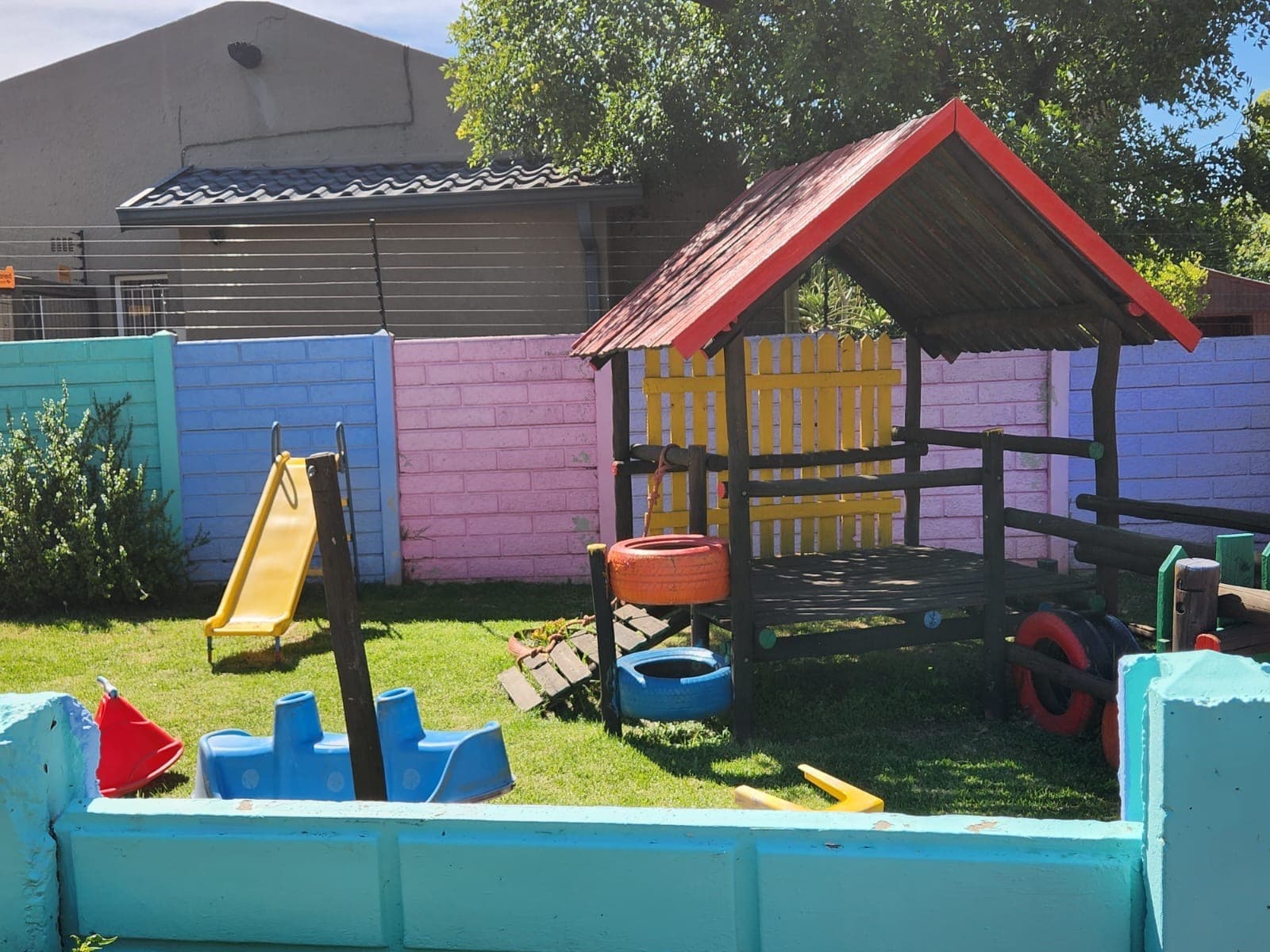 A wooden playhouse with a red roof, yellow slide and tyre seesaw on the Tiny Town lawn