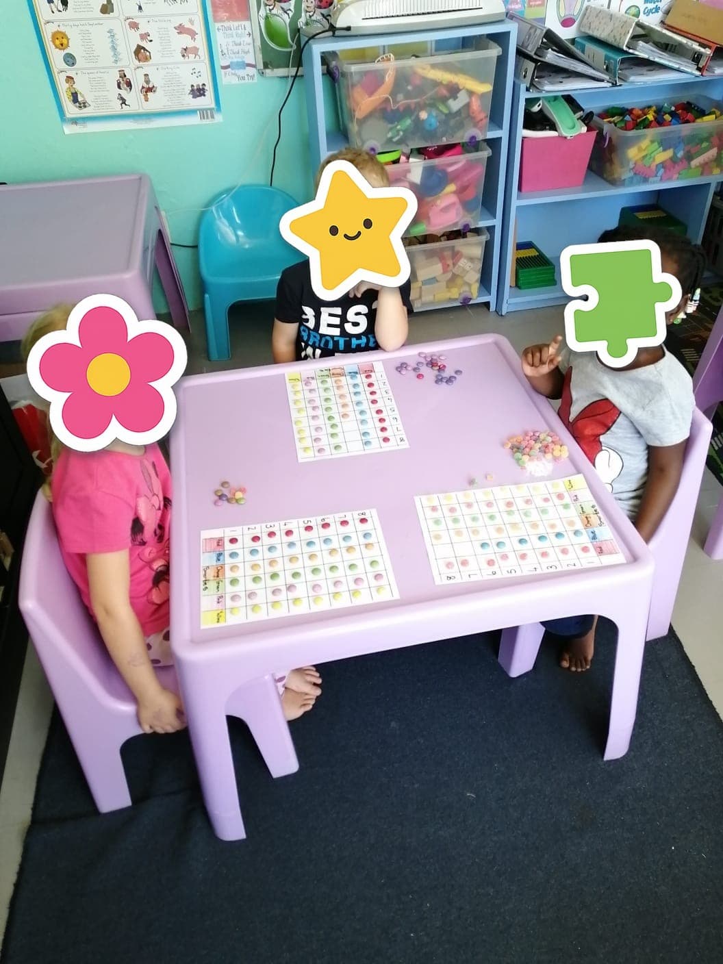 Three Tiny Town children at a pink table working through a number activity together, their faces covered with paper stickers